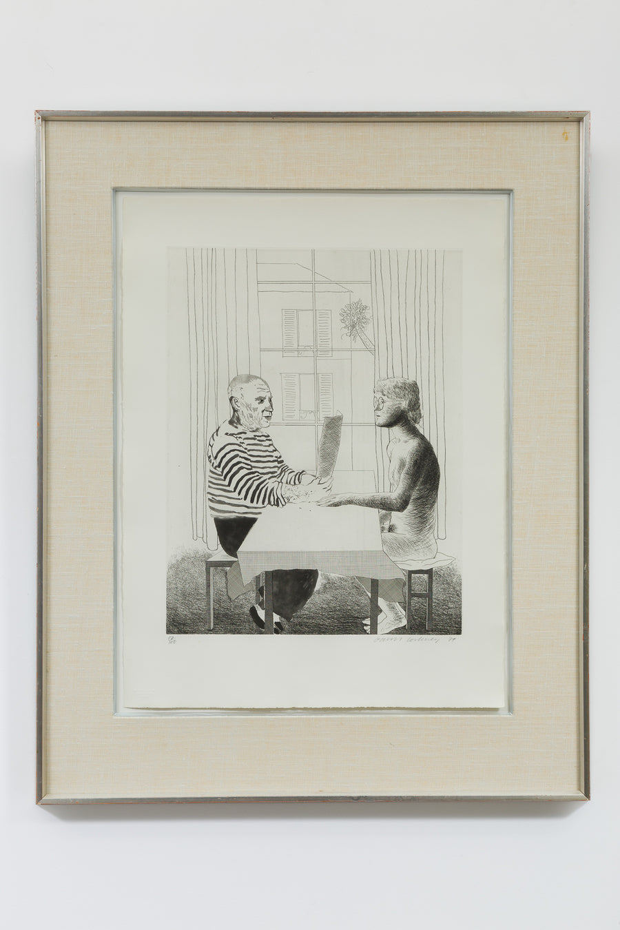 A framed etching by David Hockney, the artwork shows an artist and model seated across from each other at a table inside a private setting. The artist is wearing a striped shirt and dark pants and is holding a piece of paper in front of him. The model is a male figure with glasses, and is naked and sitting on a cushioned seat.