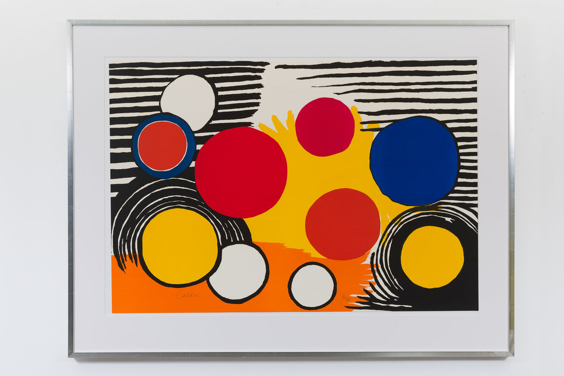 A framed print by Alexander Calder hung on a white wall. The piece includes geometric abstraction. There are several circles in the foreground, along with black strips and semi-circle lines in the background. The piece only includes primary colours.