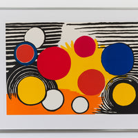 A framed print by Alexander Calder hung on a white wall. The piece includes geometric abstraction. There are several circles in the foreground, along with black strips and semi-circle lines in the background. The piece only includes primary colours.